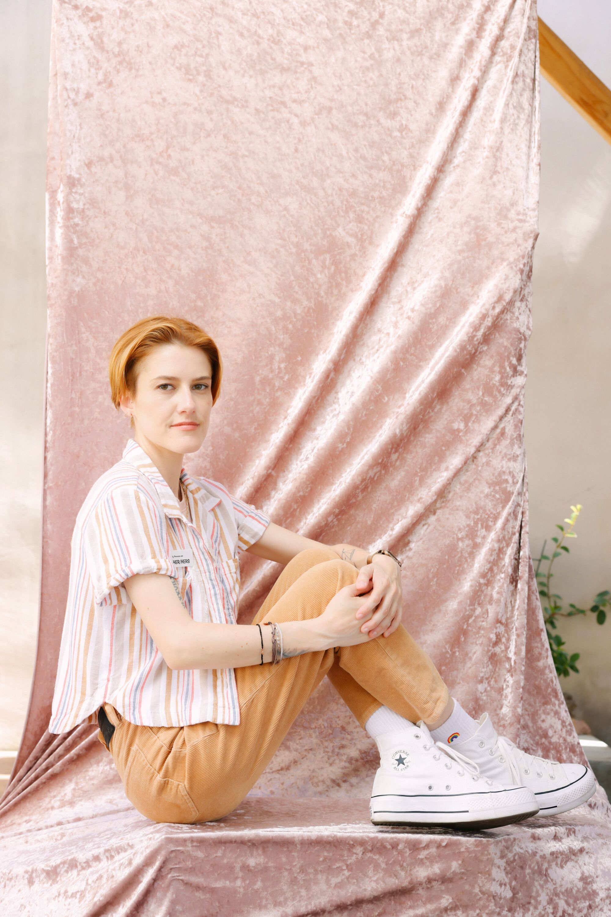 A woman with short red hair sits on a pink velvet backdrop wearing white shoes, light brown pants and a striped shirt.