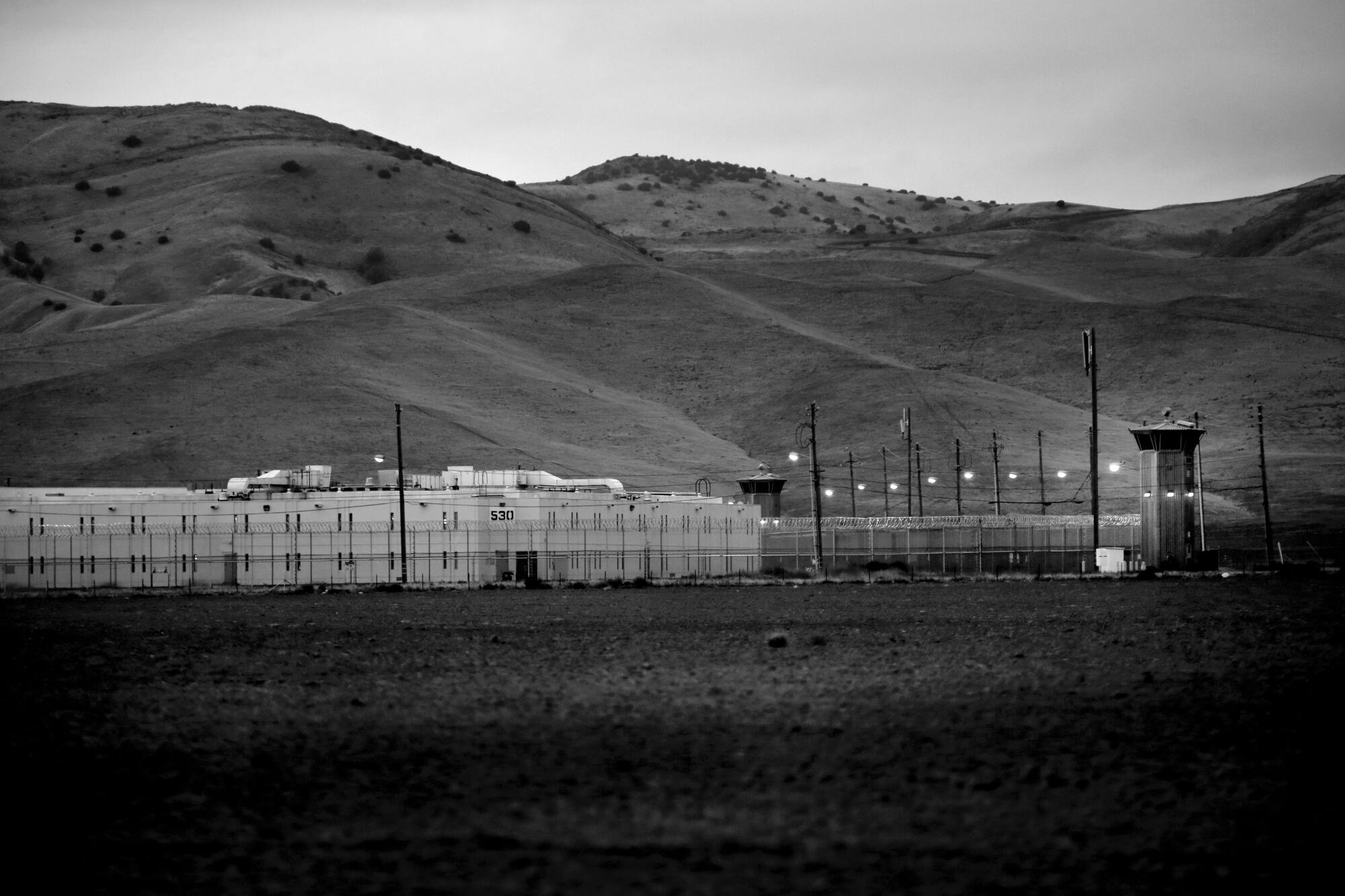 A fenced prison lies below grassy hills.