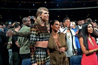 Taylor Swift, in multicolor matching set, and Kim Kardashian, in green dress,  stand arm in arm in crowd