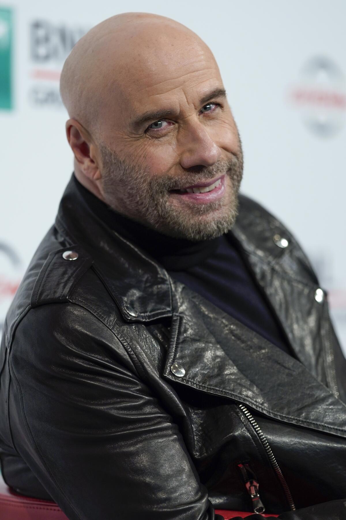 John Travolta looks to the side and smiles while wearing a black leather jacket