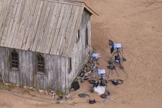 A wooden building surrounded by studio lights and equipment on a dirt surface
