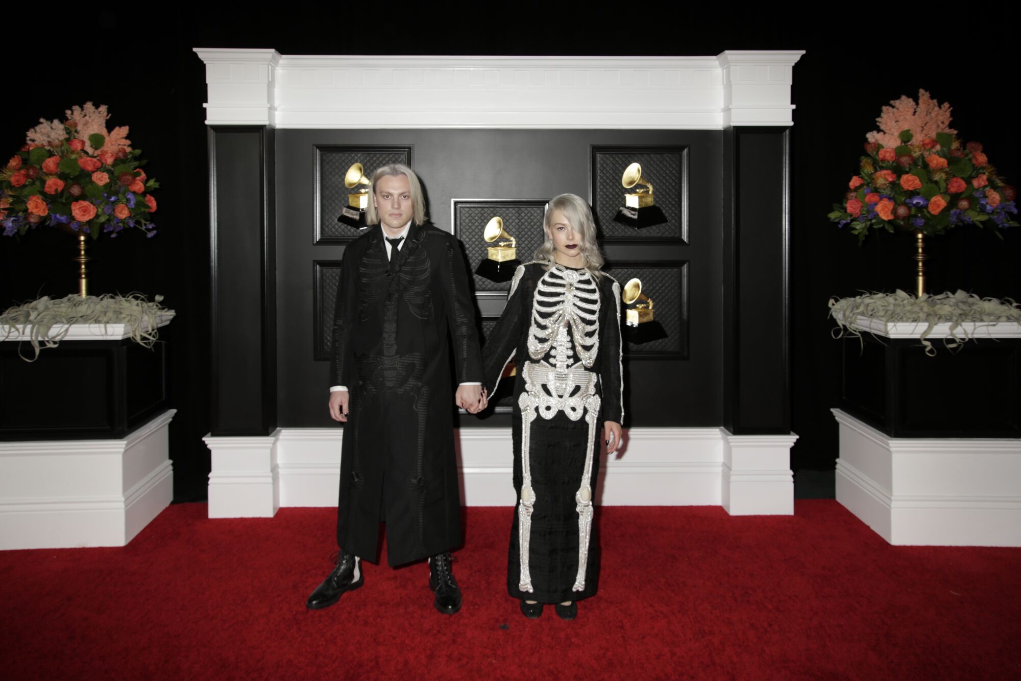 A man in black and a woman in a skeleton onesie hold hands on the red carpet.