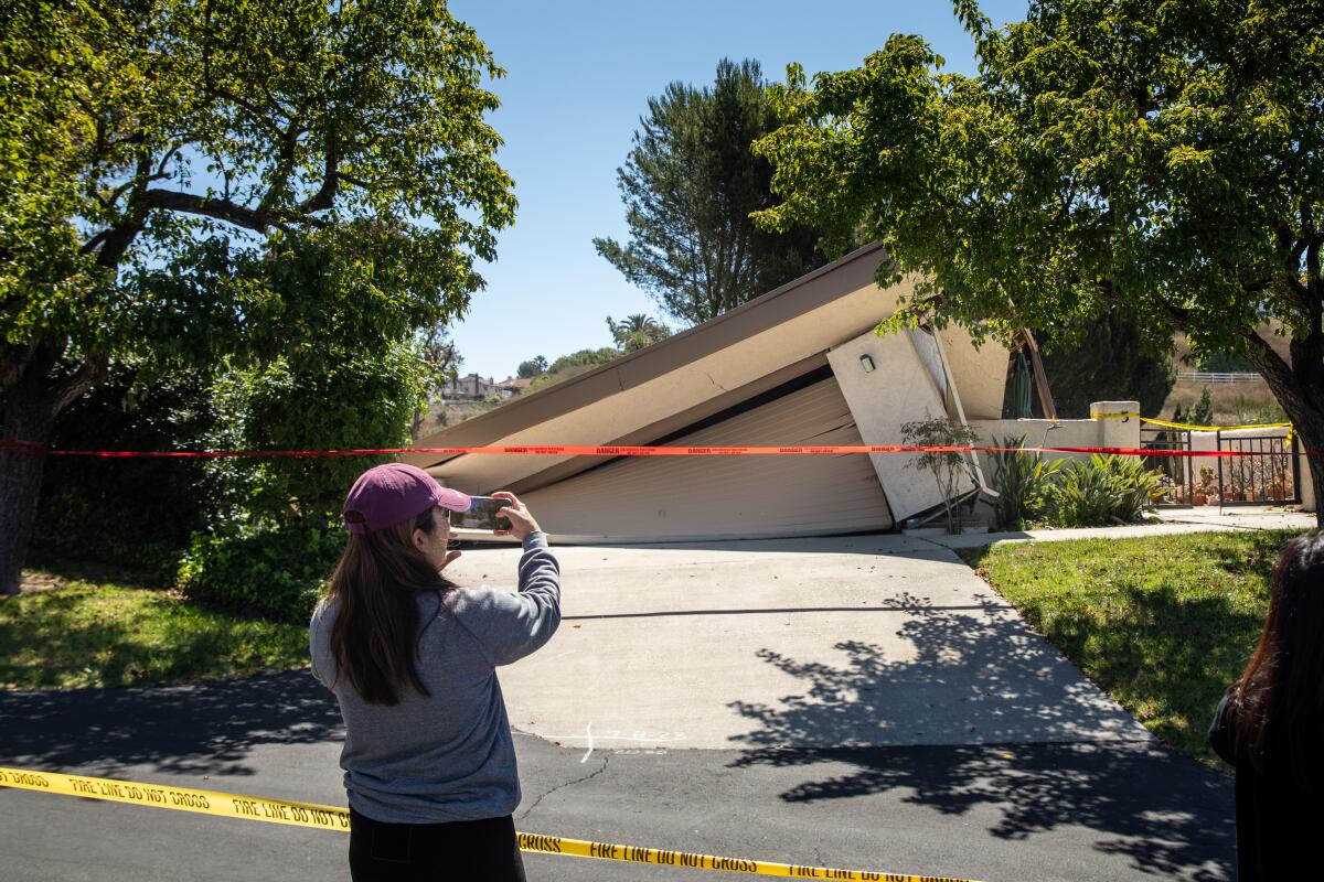 A resident of the neighborhood takes a picture of a house, which has collapsed and is sliding behind a hill and out of view.