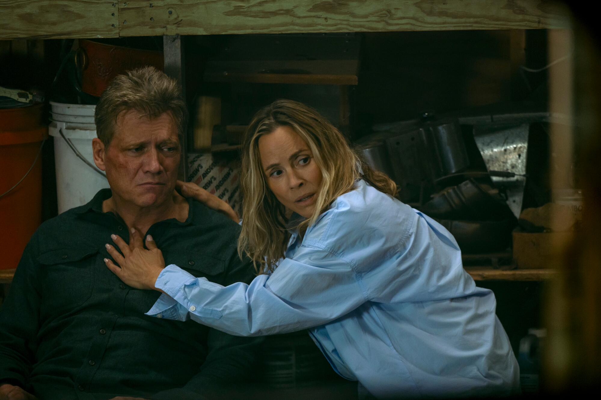 A man in a dark shirt leans against shelving as a woman in a blue shirt leans against him with a worried look on her face.