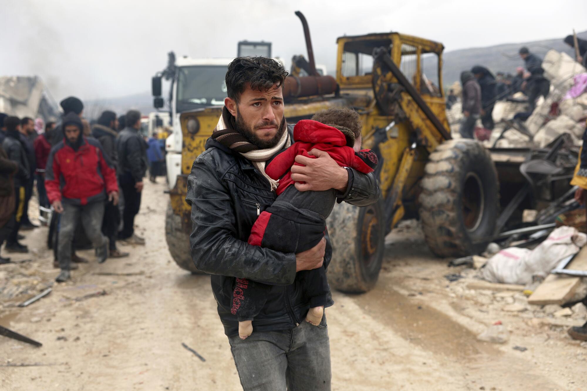 A man walks down a road between damaged buildings and machinery while he carries a young child.