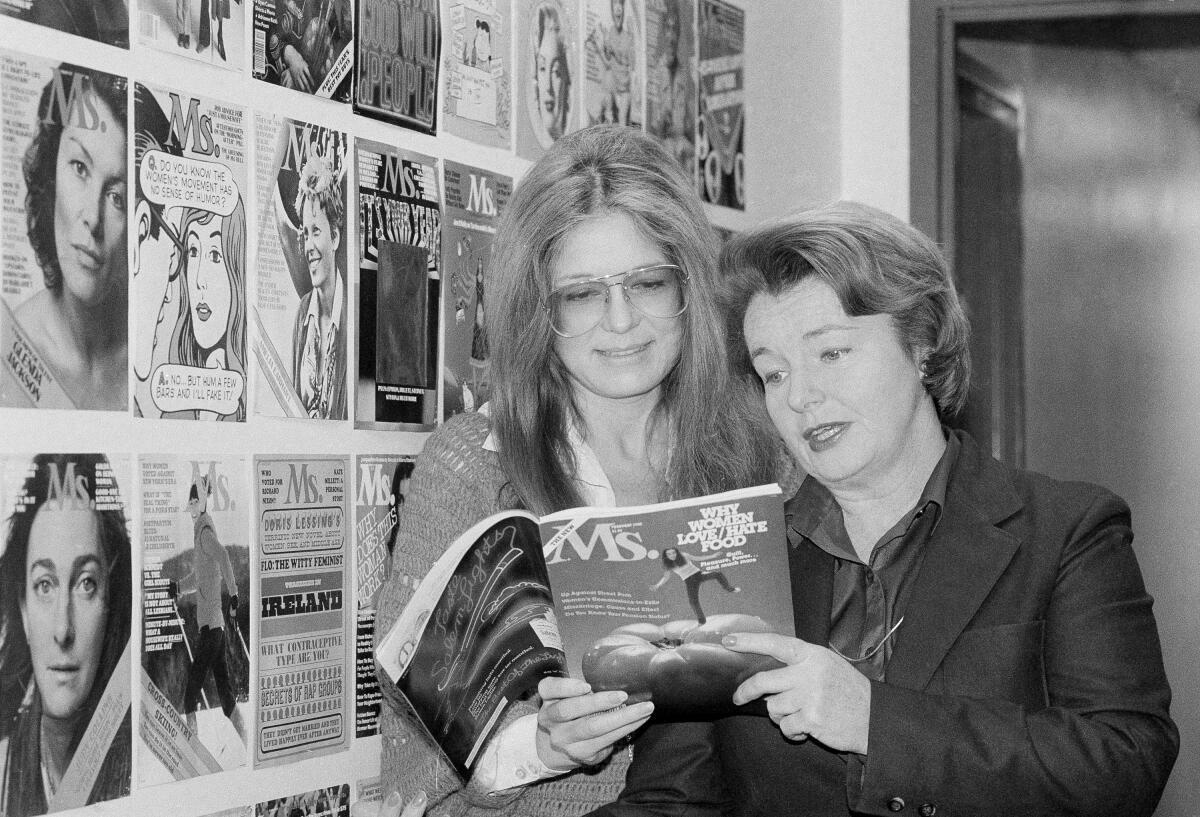 Two women looking at a magazine in front of a wall of magazine covers 