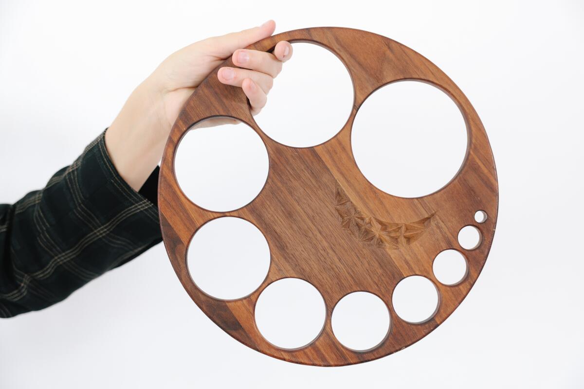 A white person's hand holds up a circular piece of wood with holes in different sizes cut out of it.