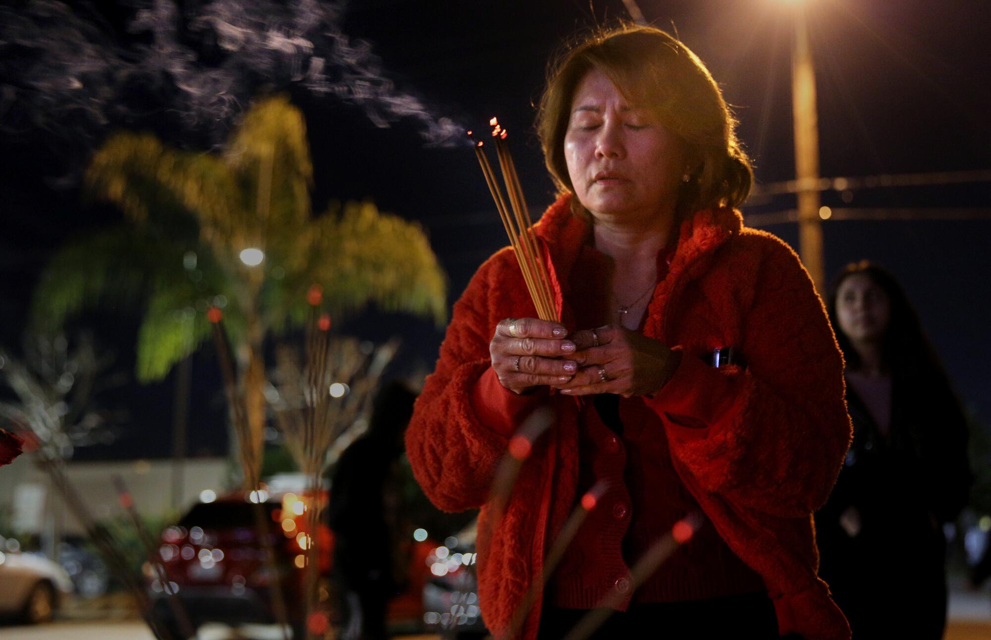 A woman holds incense sticks as she prays with her eye closed, standing outside