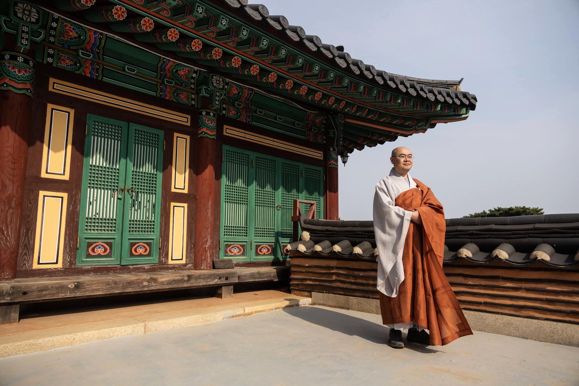 A man in white and brown monk's robes stands in front of a temple with green and brown panels 