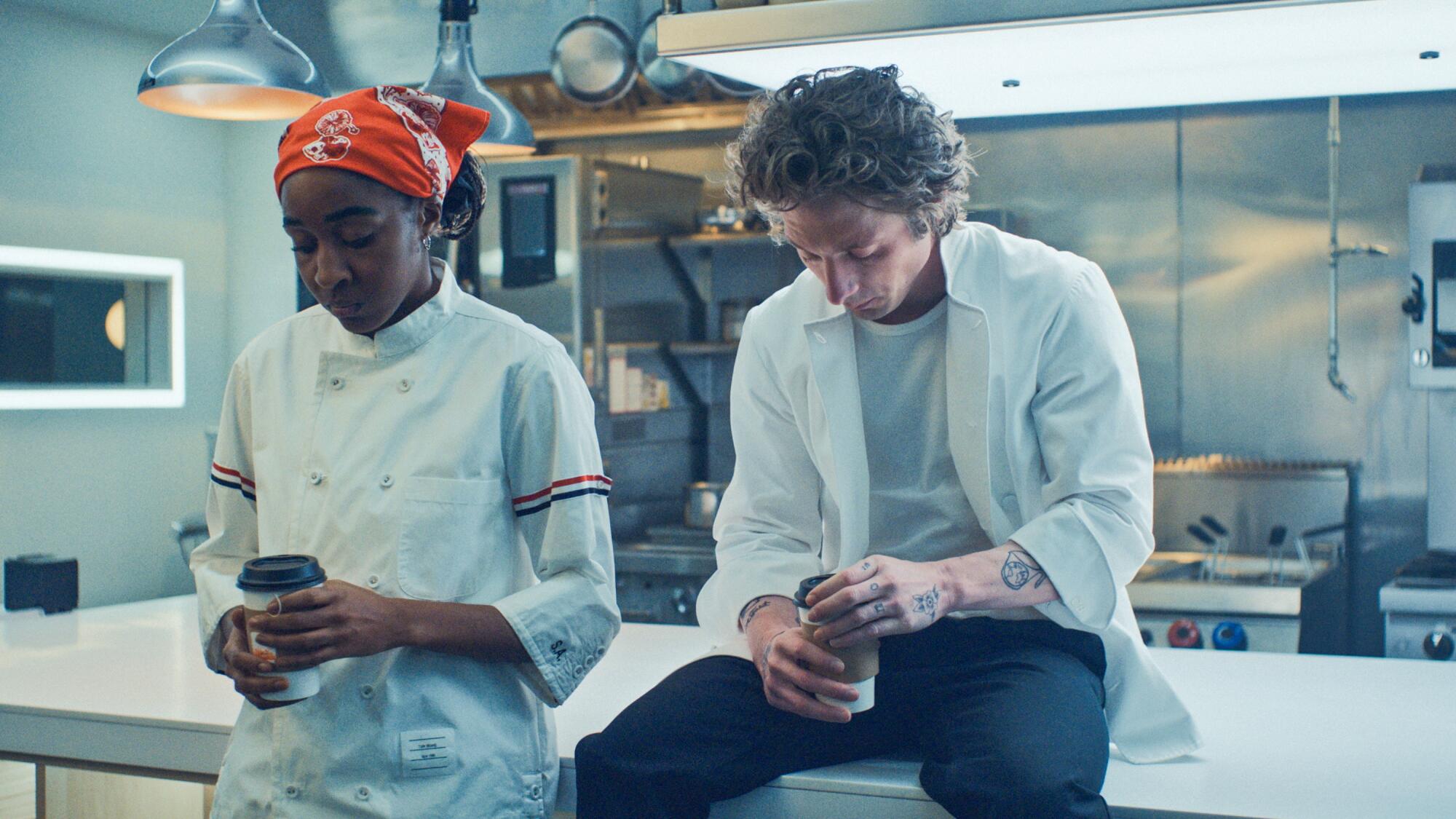 A woman in a white chef's coat and a red bandana on her head stands next to a man sitting on a prep table.