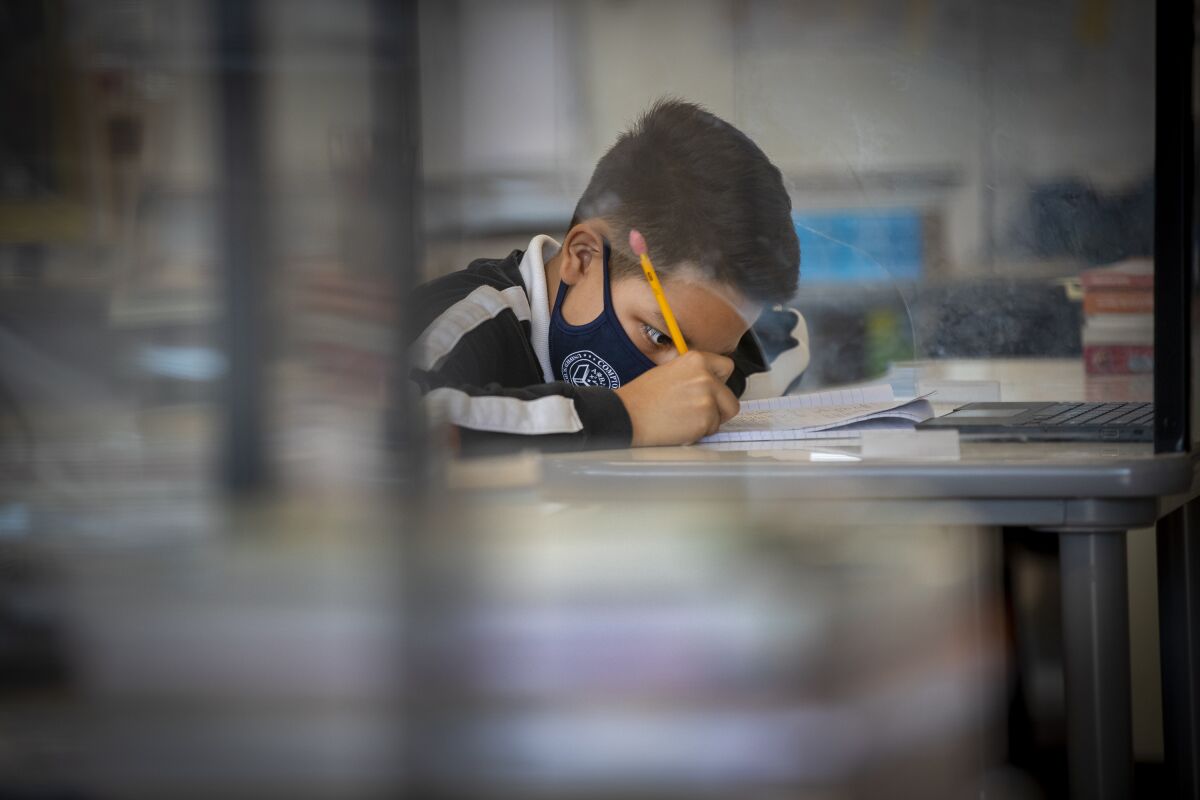 A young boy wearing a mask take notes at his classroom desk.