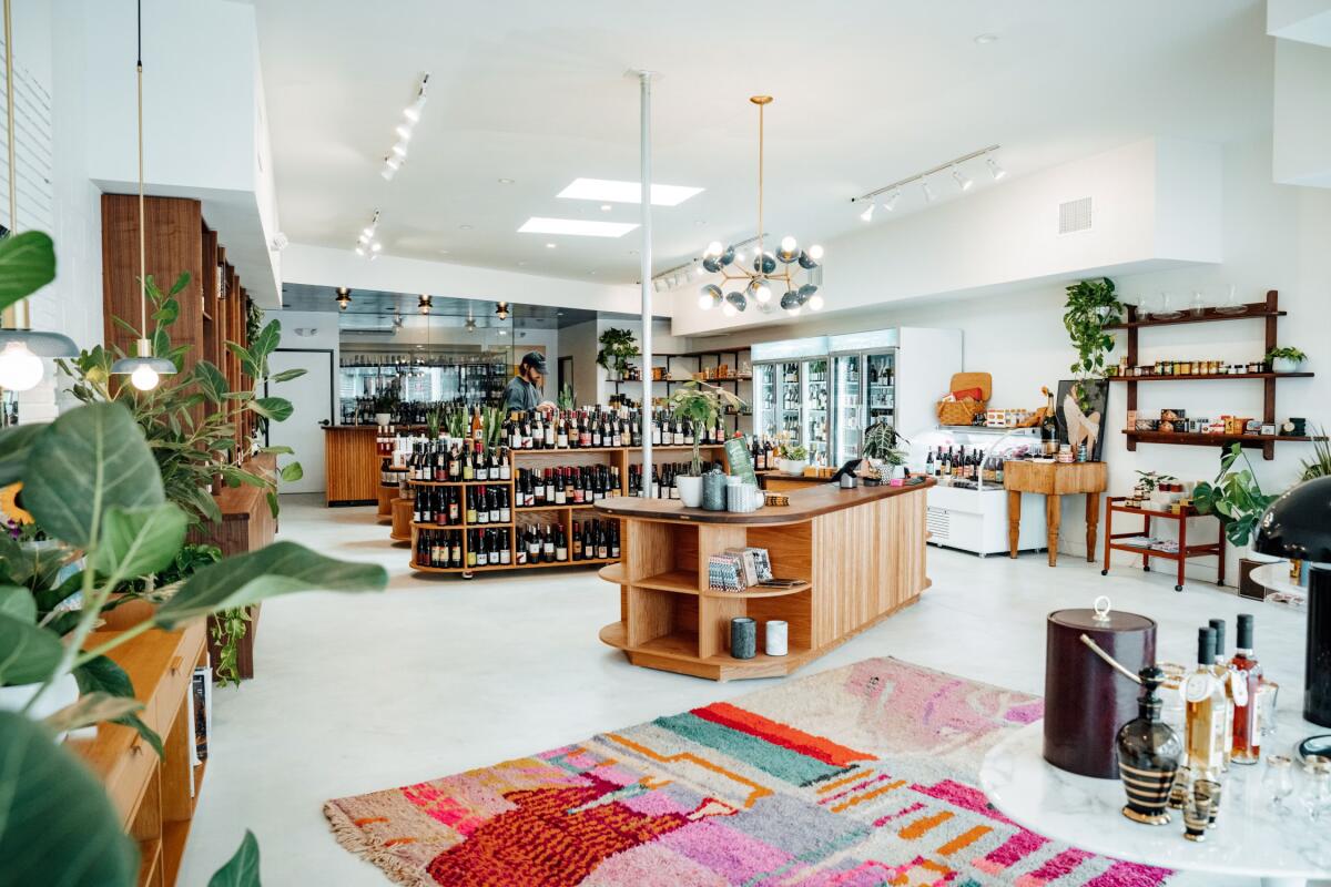 An interior featuring plants, a rug, white walls, and standalone wooden islands and wall shelves stocked with bottles of wine