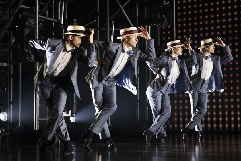 three men and one woman, all in identical suits and hats, dance and tip their hats 