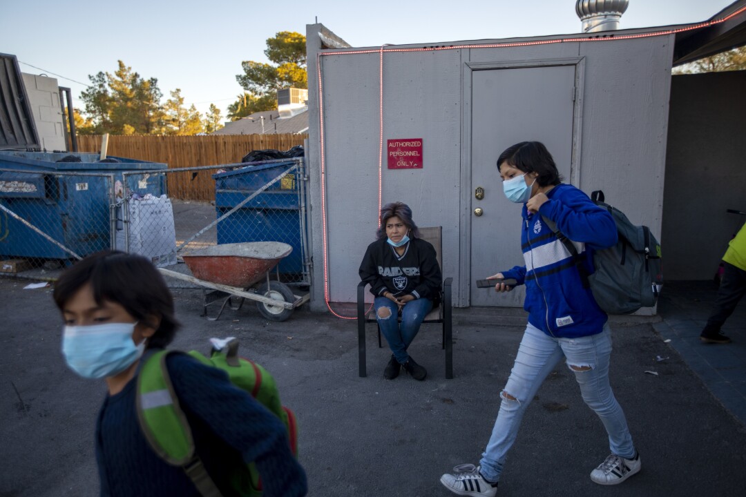 A woman sits in a chair while two young children walk past her with backpacks.