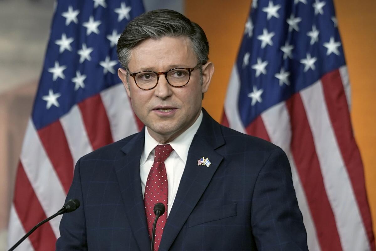 A man appears behind small microphones and in front of two American flags.