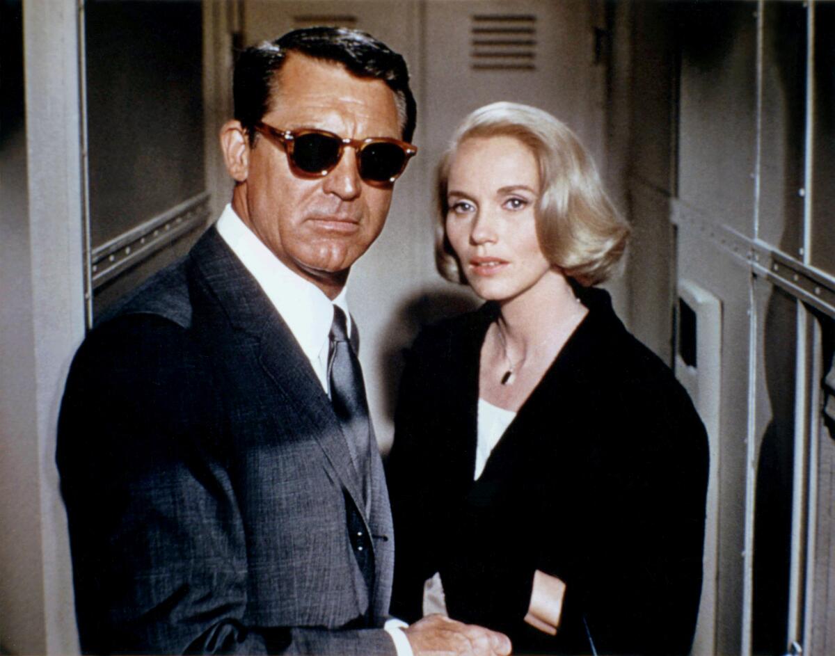 A man in sunglasses and a blond woman in a dark suit face each other and look toward the photographer.