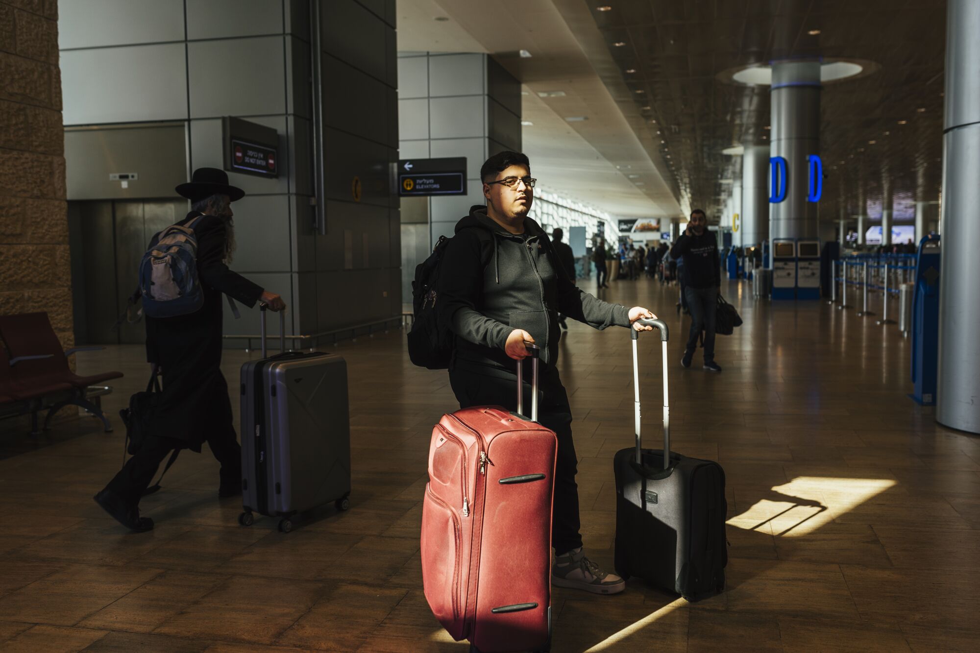 A man with dark hair and glasses, wearing a backpack, holds the handles of a red suitcase and a smaller, black one
