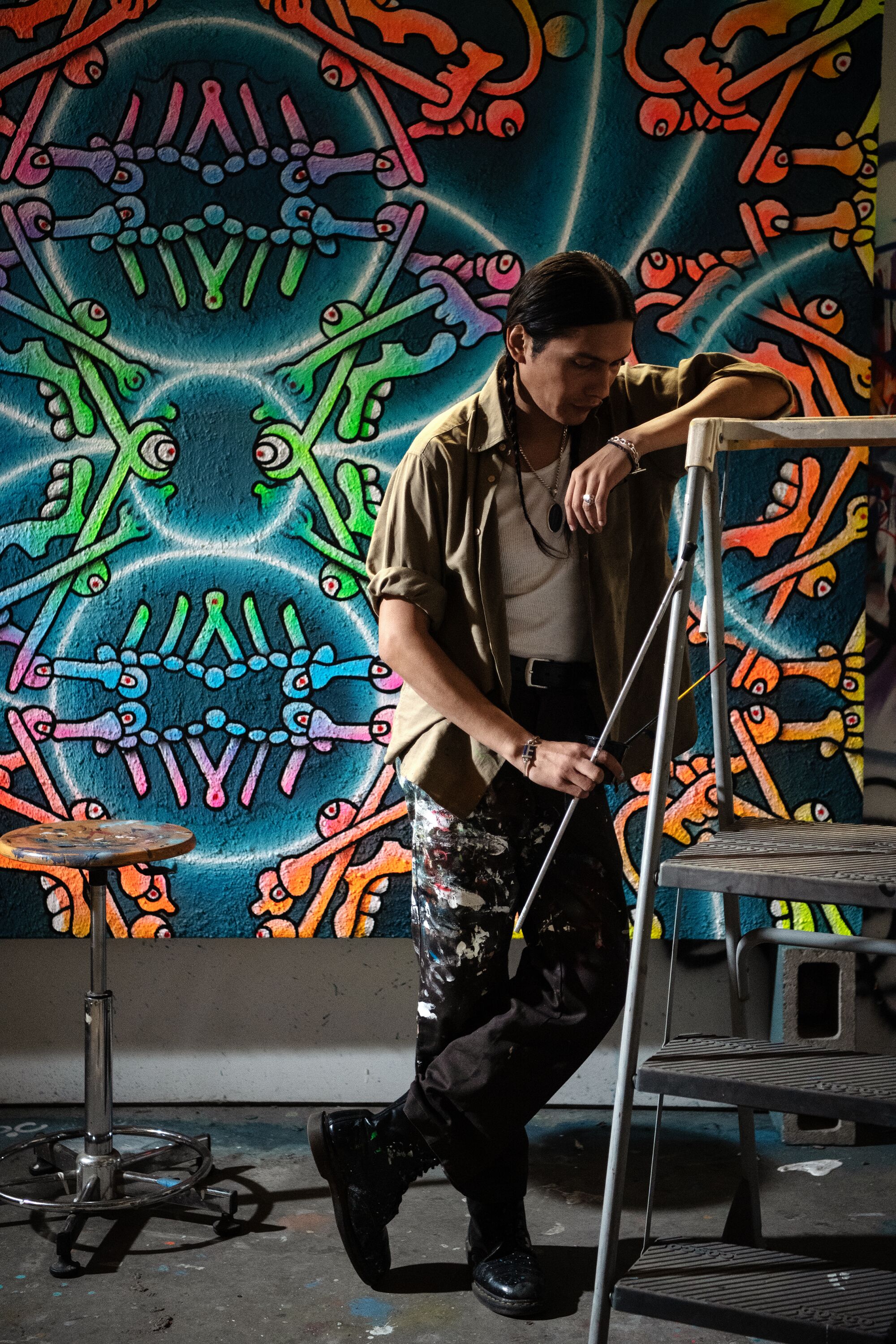 ozzie juarez leans on a stepladder, one of his vibrant paintings in the background