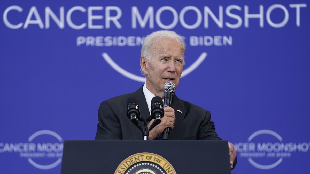 A gray-haired man in a dark suit speaks holding a microphone, with a purple background and the words Cancer Moonshot