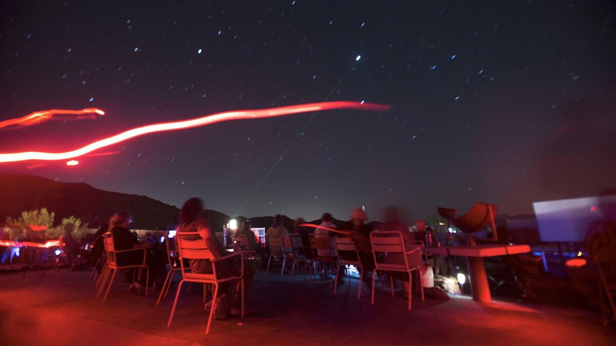 A long camera exposure captures guests staring at twinkling stars and a giant laser beam pointing to Europa, Jupiter's moon, at the Astronomy Arts Theater in Joshua Tree, Calif. The red light on the left is a flashlight used by guides.