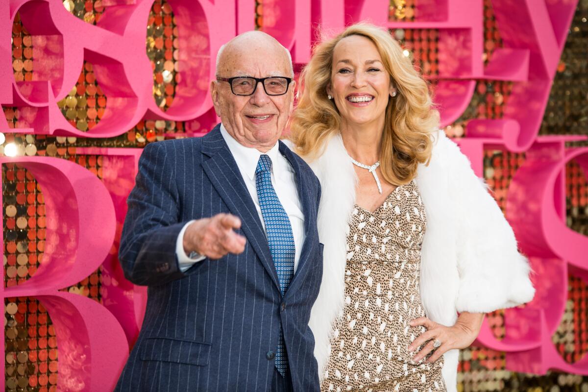 An elderly man with glasses and a suit poses for pictures alongside a blonde woman in a sparkling gown and a white coat. 