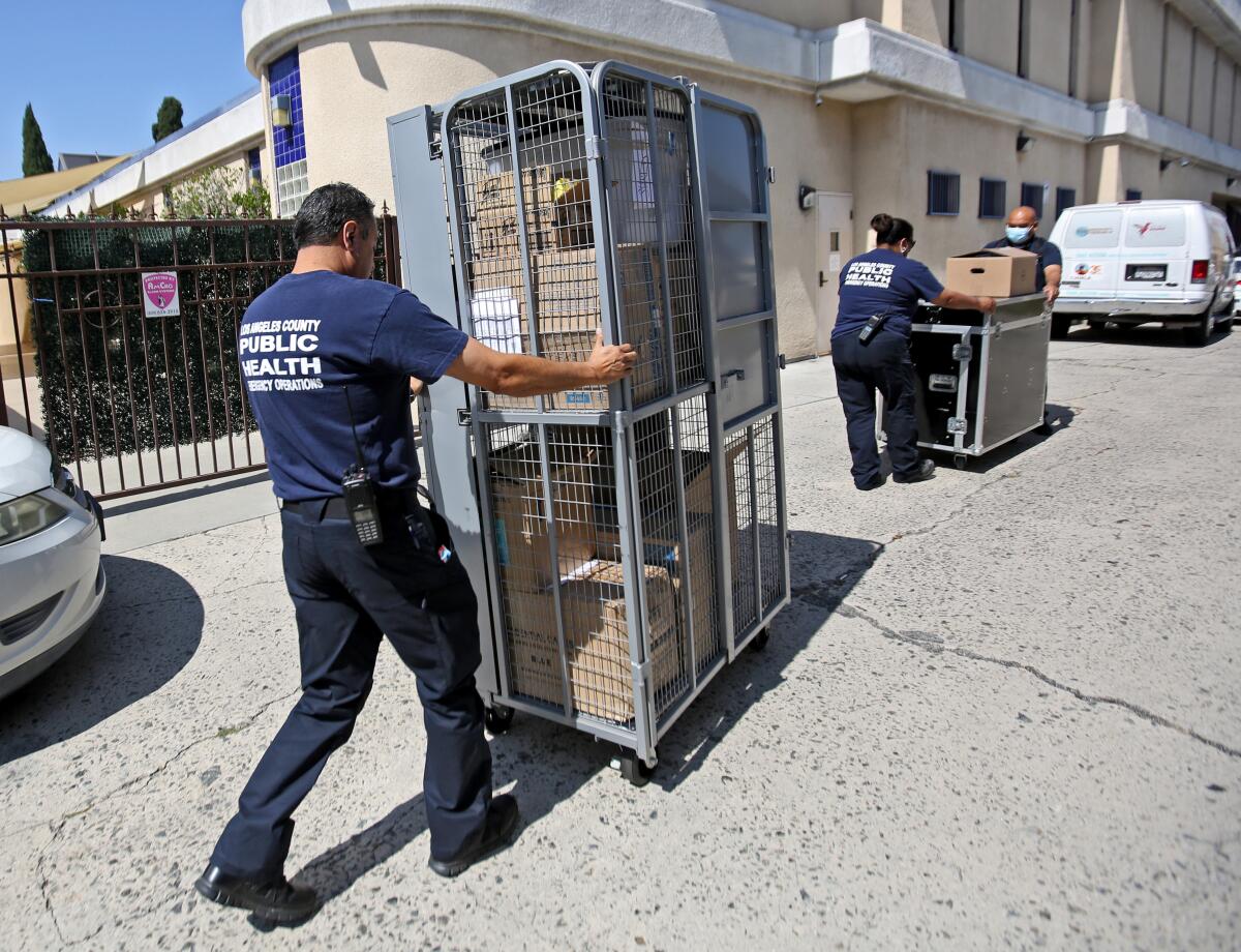 Los Angeles County Public Health workers push large metal carts outside a building  