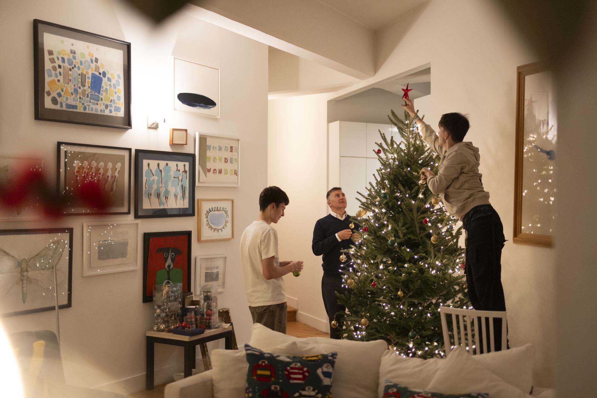 A young man stands on a chair to decorate a Christmas tree as his father and brother stand nearby.