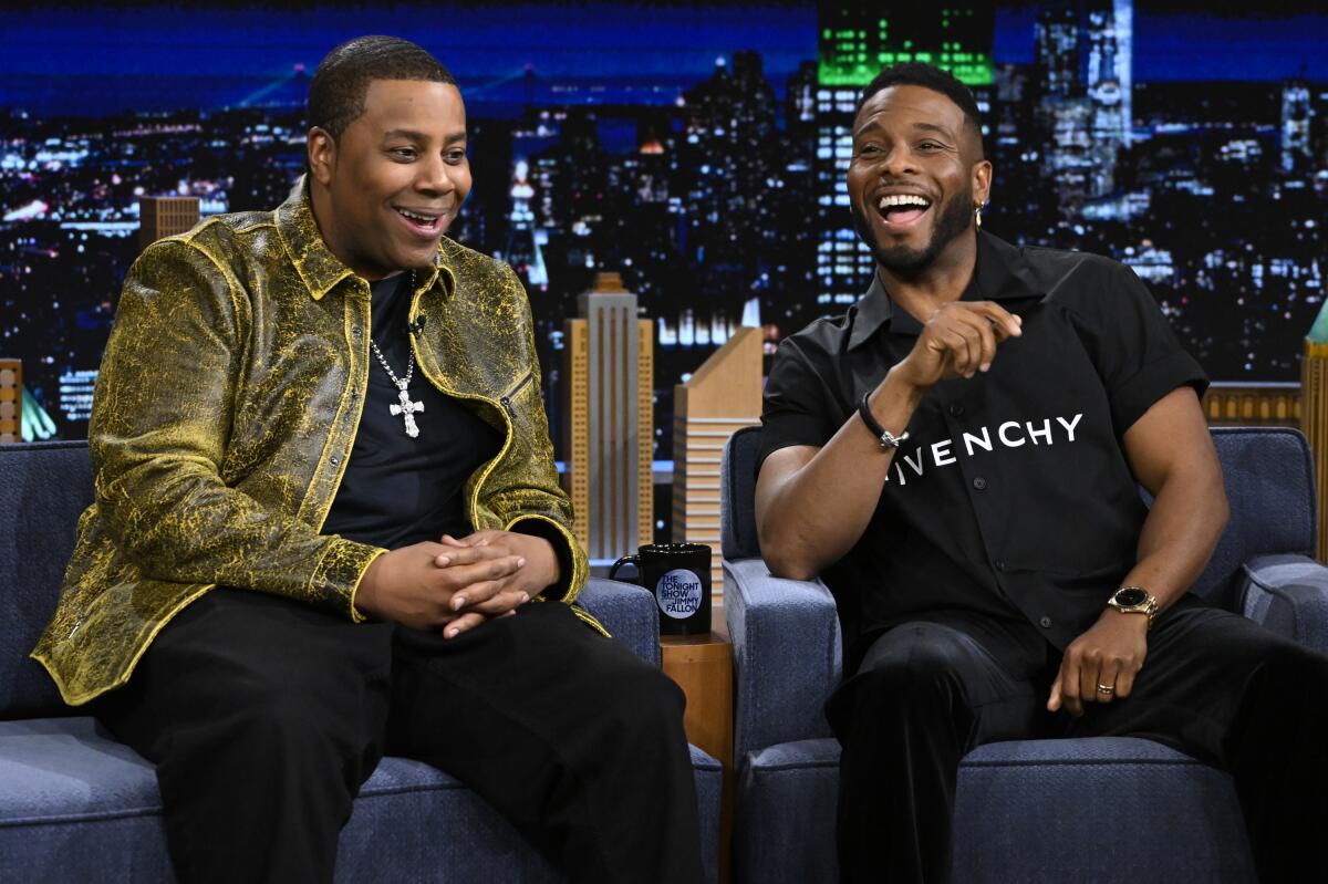 Two actors sit talking and laughing on the set of a late-night TV talk show.