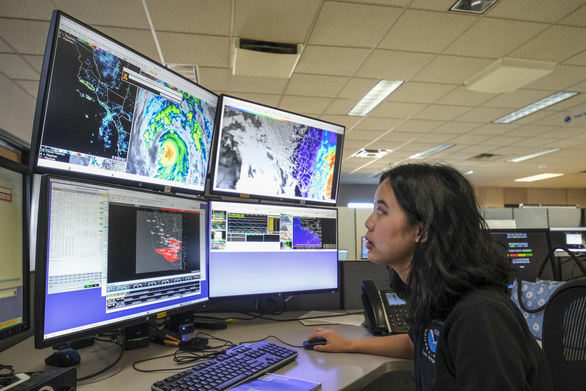A woman sits in front of an array of computer monitors showing weather information.