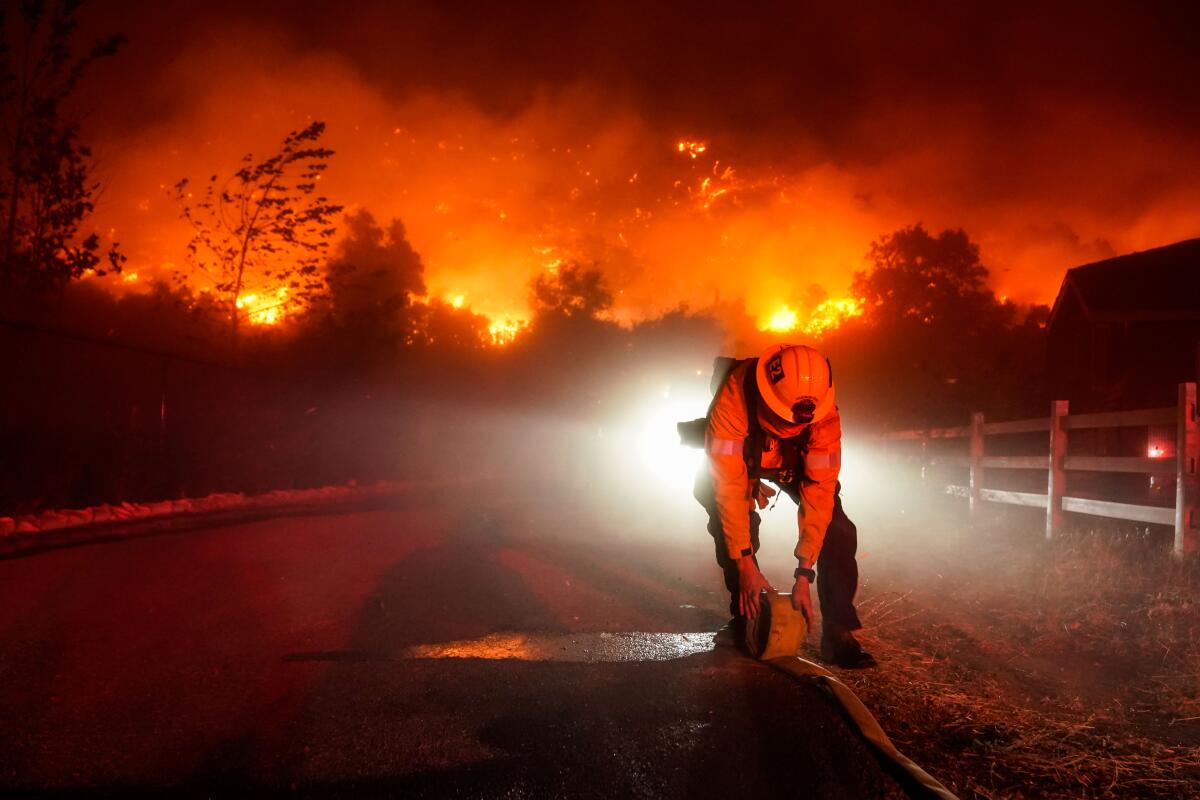 A helmeted firefighter bends over a rolled-up firehose on a roadway as flames glow in the background.