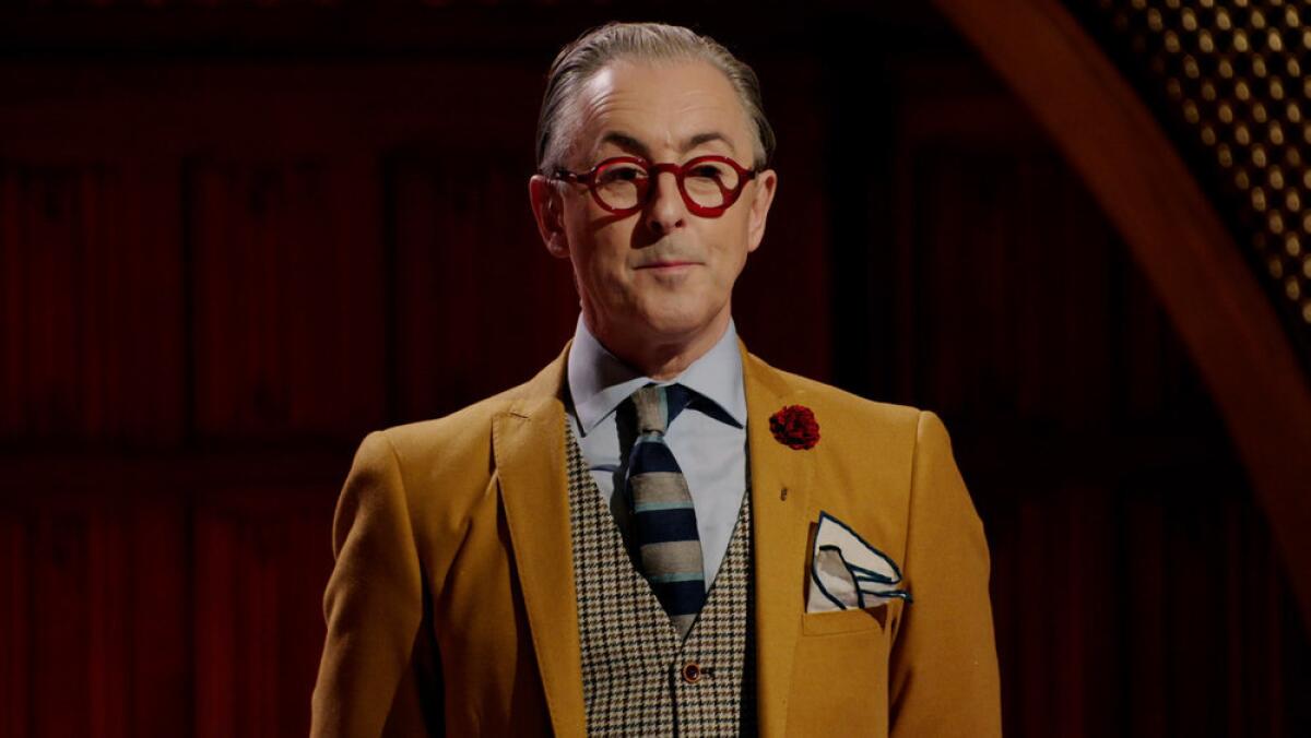 A man in an ochre suit and thick-rimmed round specs.