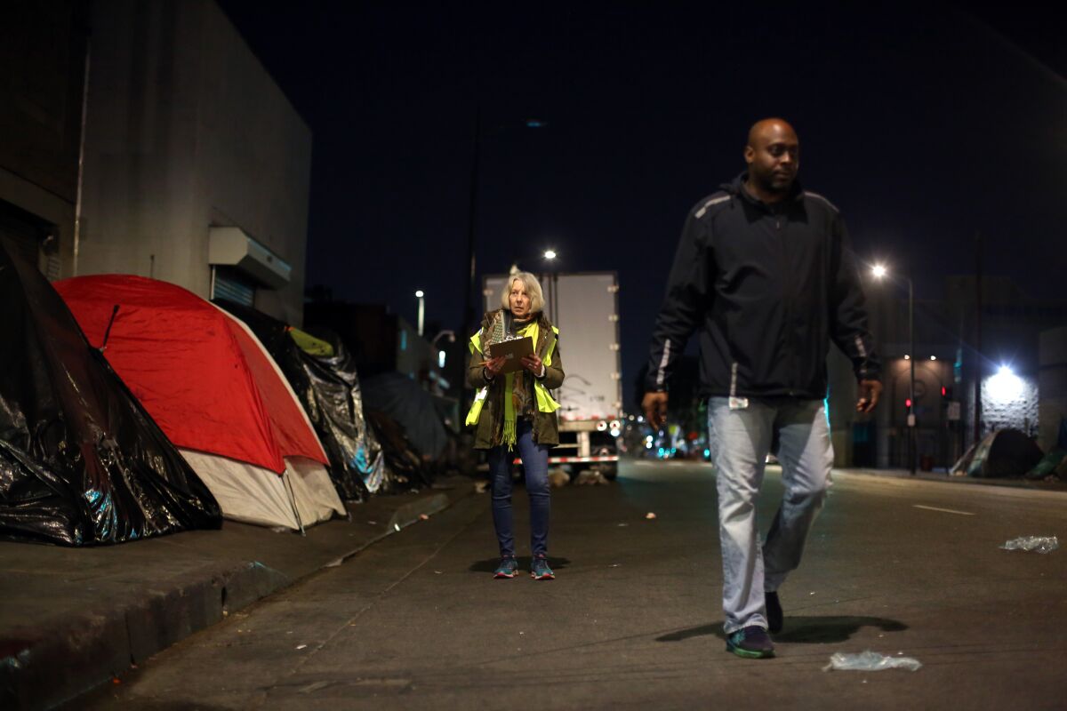 Two people walk past tents on the sidewalk at night.