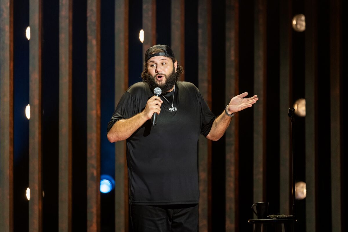 A bearded stand-up comedian in a black T shirt, backwards cap and silver chain