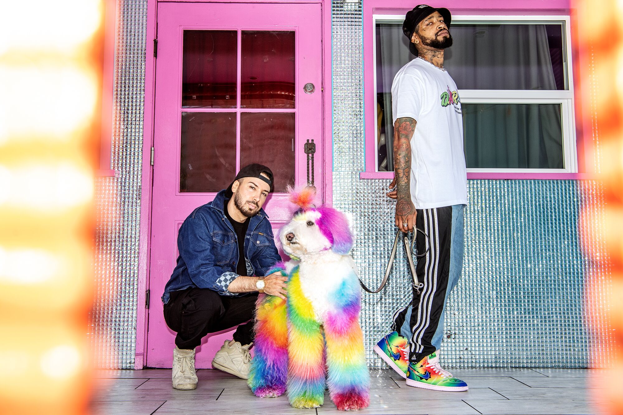 A man crouches down with a tie-dyed dog while another man stands holding a leash