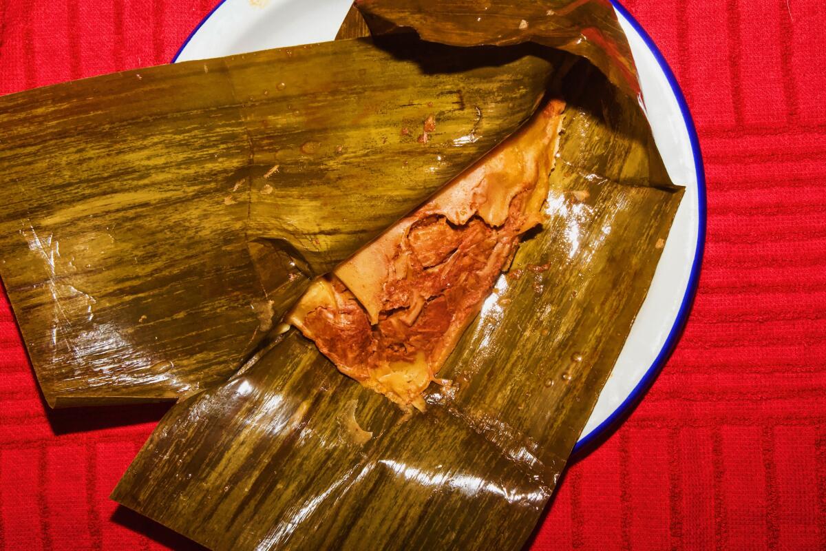 An overhead photo of a partially unwrapped rojo tamale on a plate against a red background.