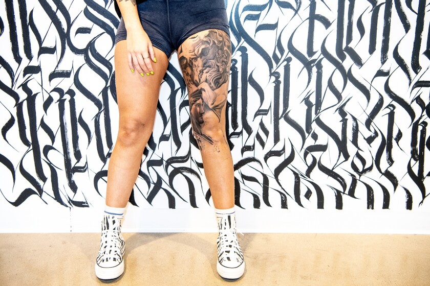 A woman's legs with tattoos on one thigh against a black-and-white background of stylized calligraphy