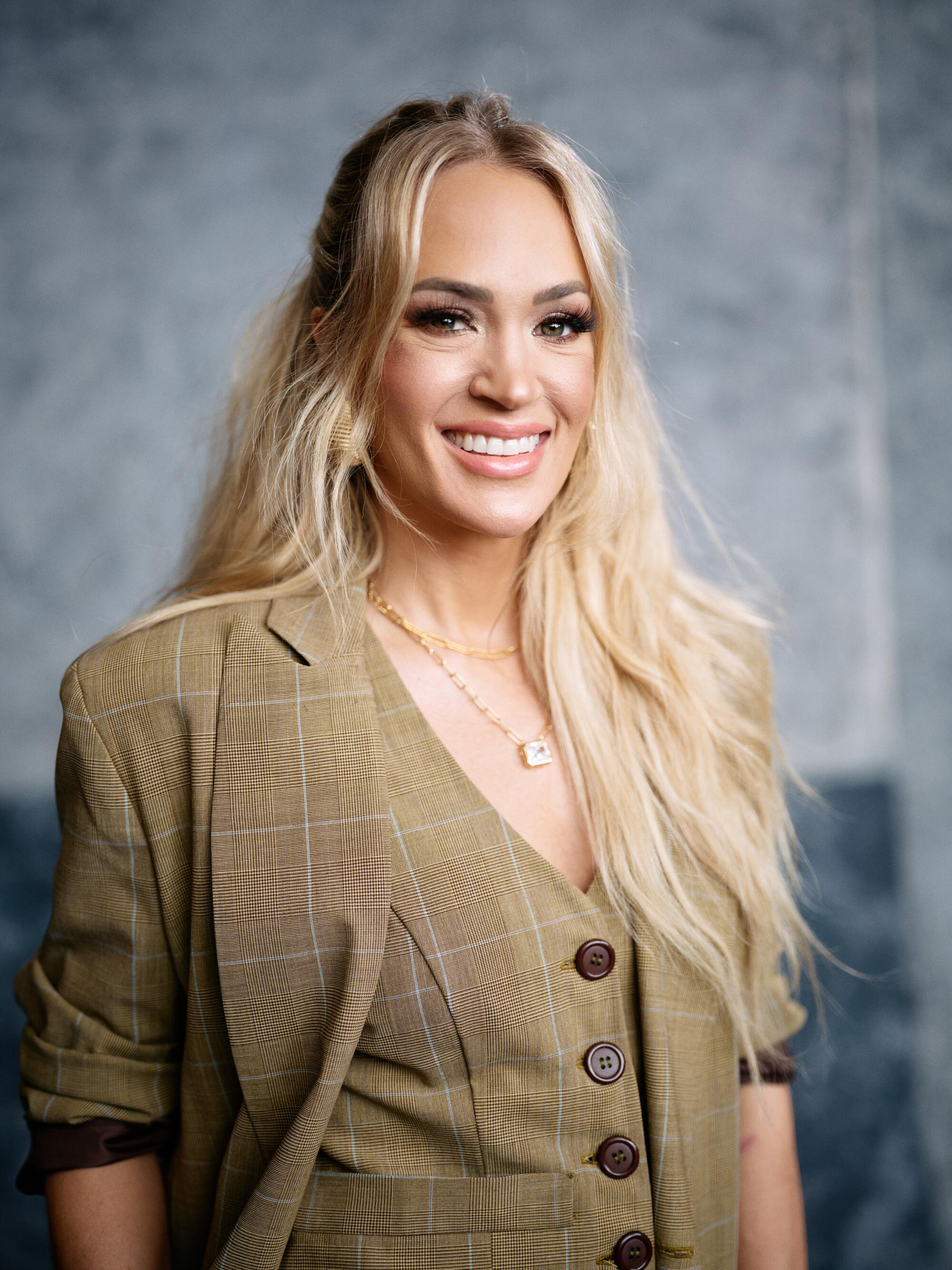 A woman with long blond hair in a tan plaid suit smiles.