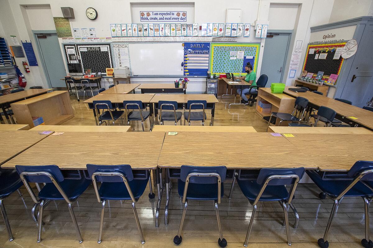 A teacher sits at a desk with an open laptop in a classroom of empty desks.