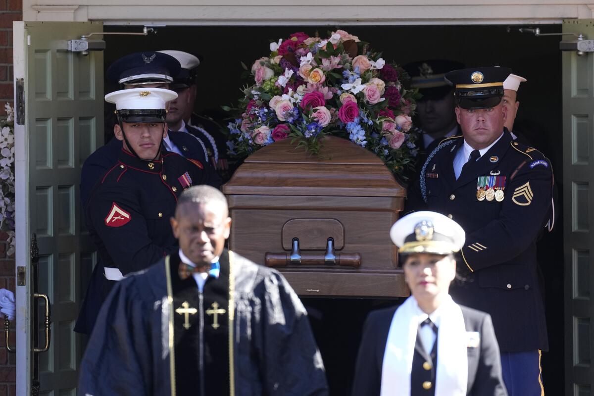 A man wearing a dark robe and a woman wearing a uniform stand in front of a coffin carried by men in uniforms.