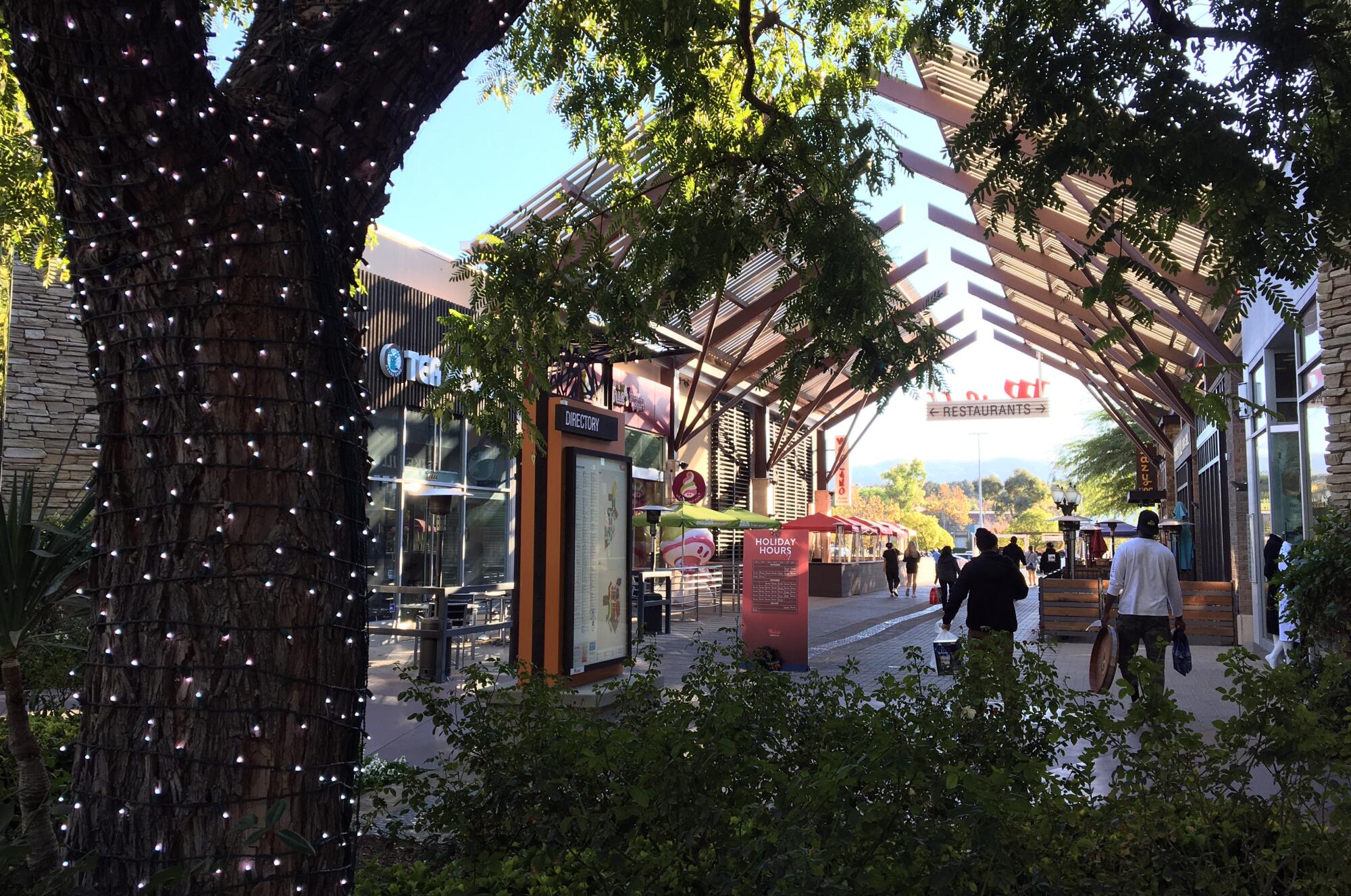 A few people carry bags at an outdoor mall courtyard, where a tree is covered in holiday lights.