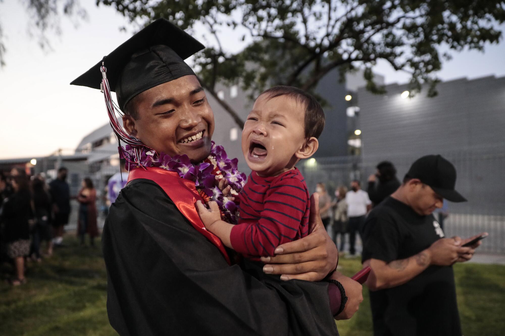 Johnny Sen, wearing a graduation cap and gown, holds a crying child.  