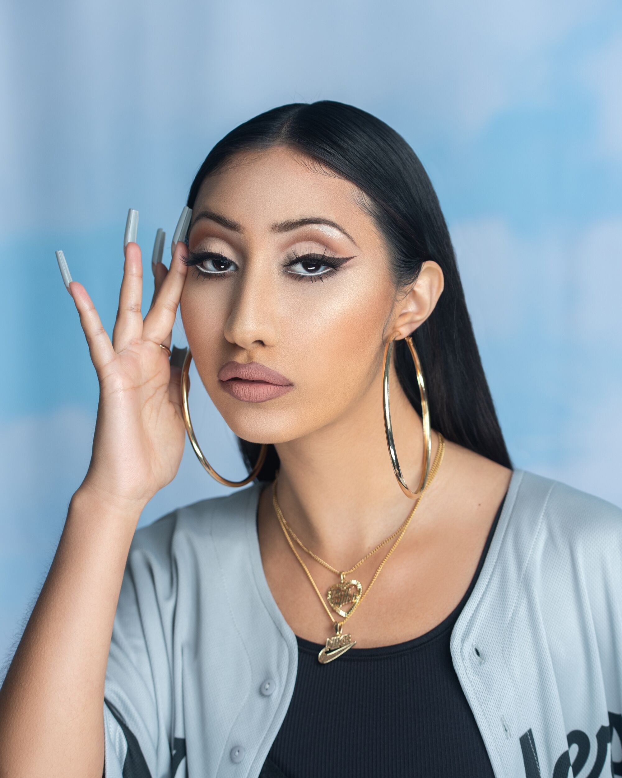 a brown model with straight black hair and Sweet Street winged eyeliner, wearing large gold hoops and layered gold necklaces