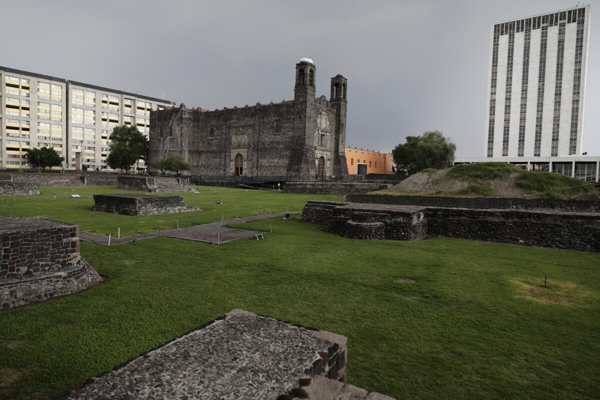 A Spanish colonial church, center, is surrounded by stone ruins and lawn, and is flanked by modern buildings 