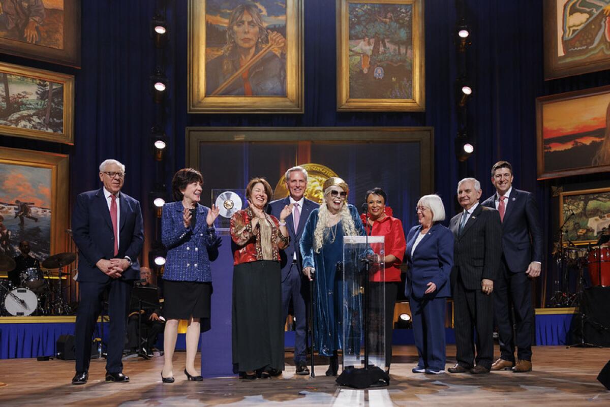 A group of men and women in suits surrounds a gray-haired woman wearing a gold beret onstage.