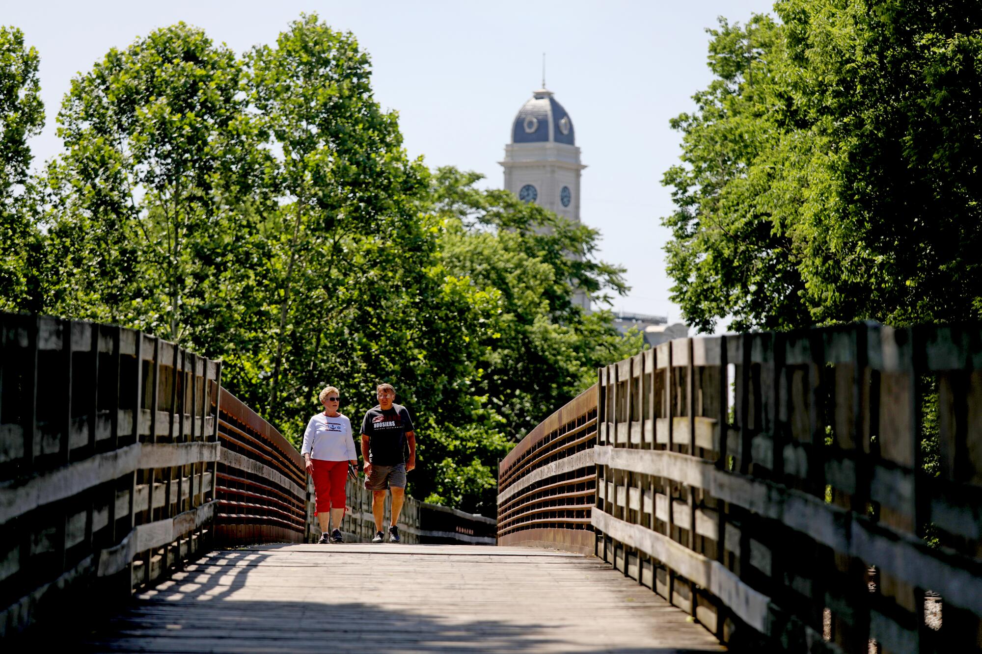 A couple walks across a footbridge in a wooded area with a cupola of a building in the distant background