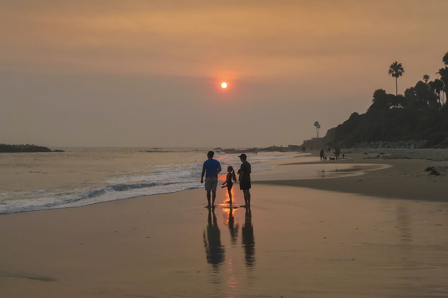 Beachgoers walk along the shoreline in Laguna Beach beneath a hazy sky.