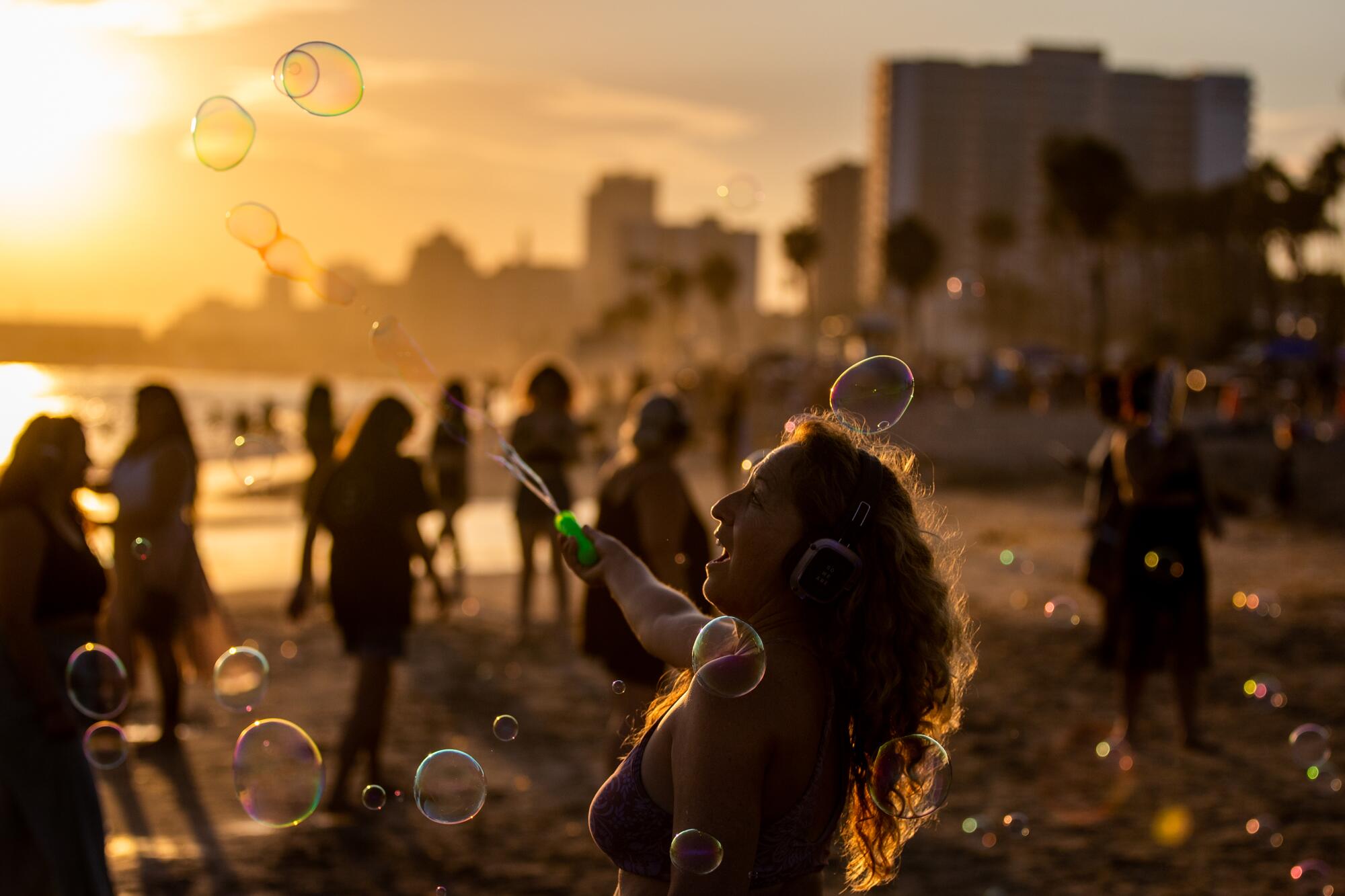 A sunset image of a woman wearing headphones, dancing on the beach and creating bubbles with a bubble wand.