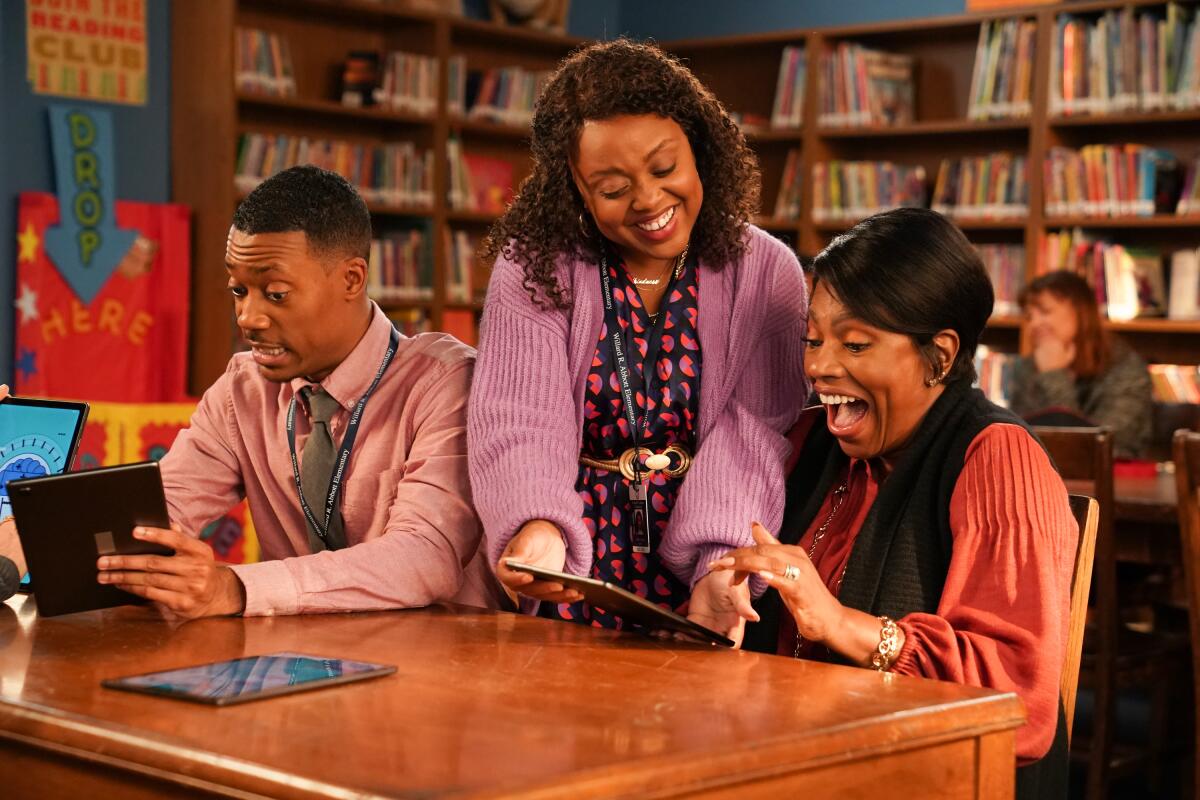 Two women at a table laugh at something on a tablet while a man next to them tries to read his own tablet.