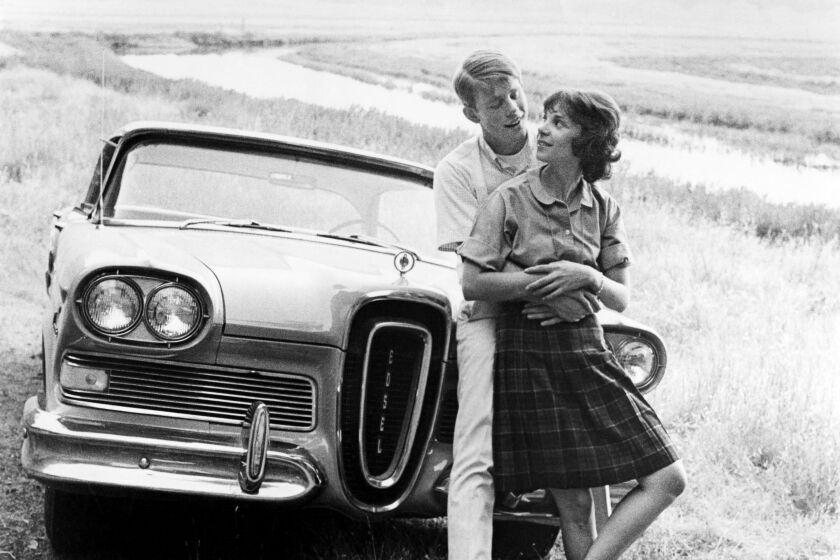 A black and white photo of a man holding a woman in front of a vintage car