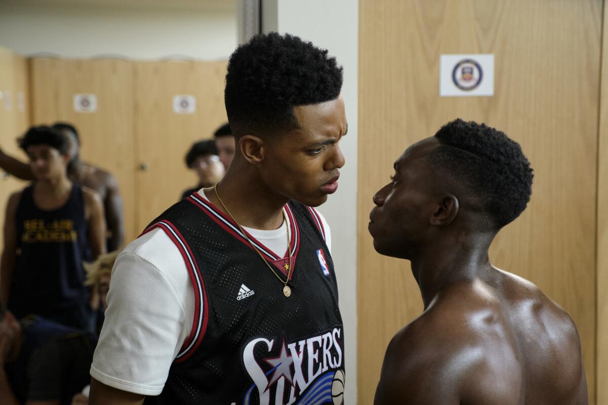 A tall man in a Sixers jersey looks at a shirtless man.